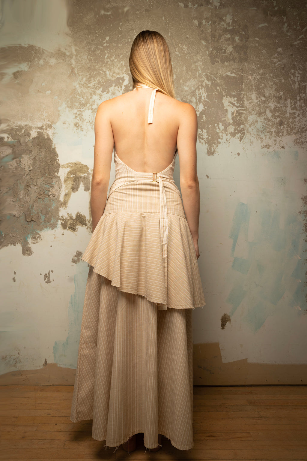 A woman stands with her back to the camera wearing a beige and white vertically striped halter dress with a ruffled asymmetrical hem, posed in front of a rustic, textured plaster wall.