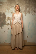 A woman stands barefoot against a distressed, textured wall wearing a cream halter vest with a buttoned waist and a long beige tiered skirt.