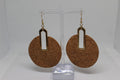 A pair of large circular cork earrings with gold-toned metal accents and hooks, displayed on a clear acrylic stand against a white background.