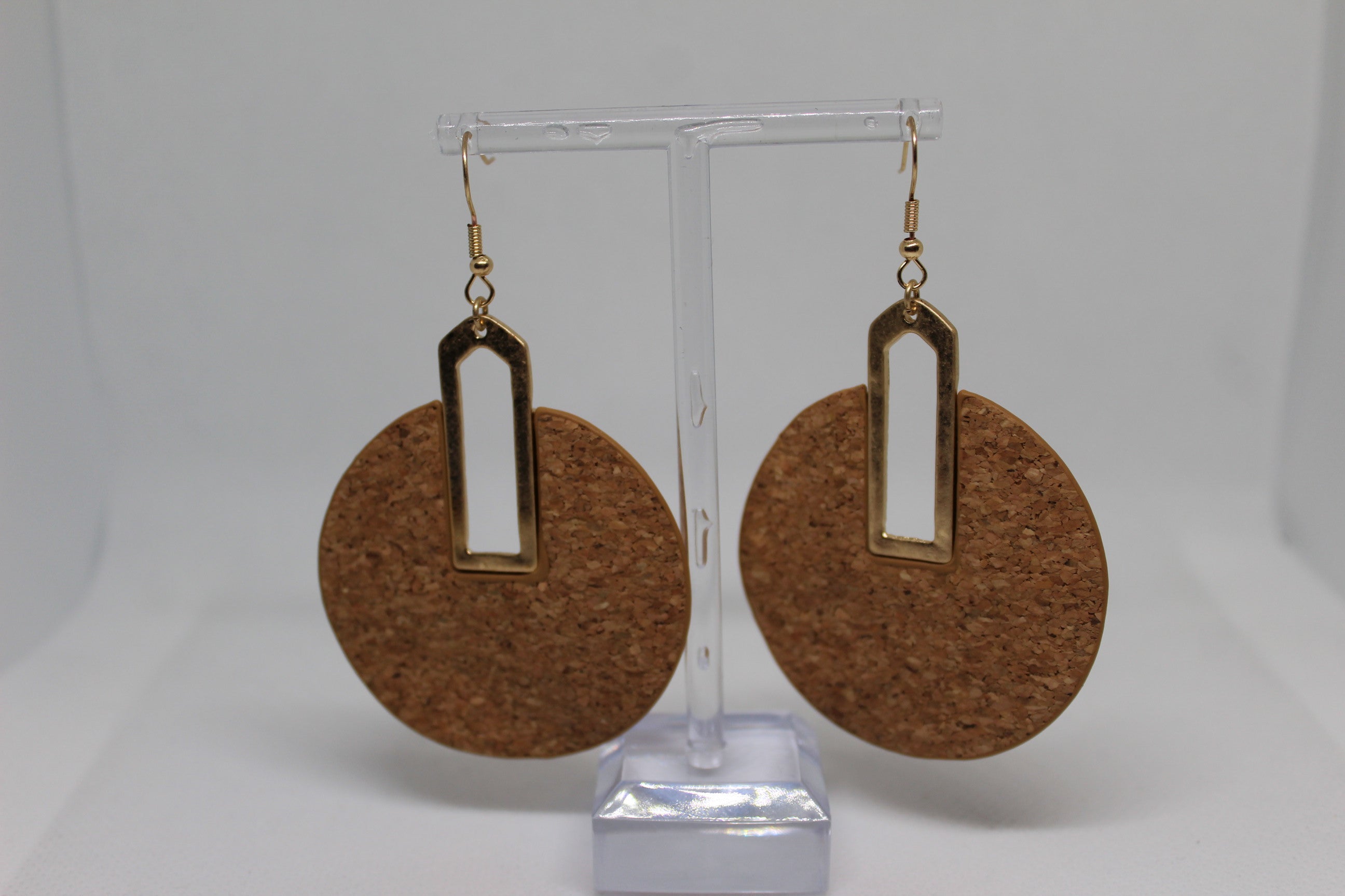 A pair of large circular cork earrings with gold-toned metal accents and hooks, displayed on a clear acrylic stand against a white background.