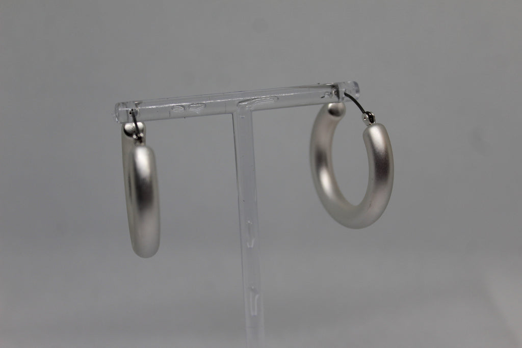A pair of large, polished silver hoop earrings hanging from a clear plastic display stand against a neutral gray background.