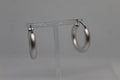 A pair of large, polished silver hoop earrings hanging from a clear plastic display stand against a neutral gray background.