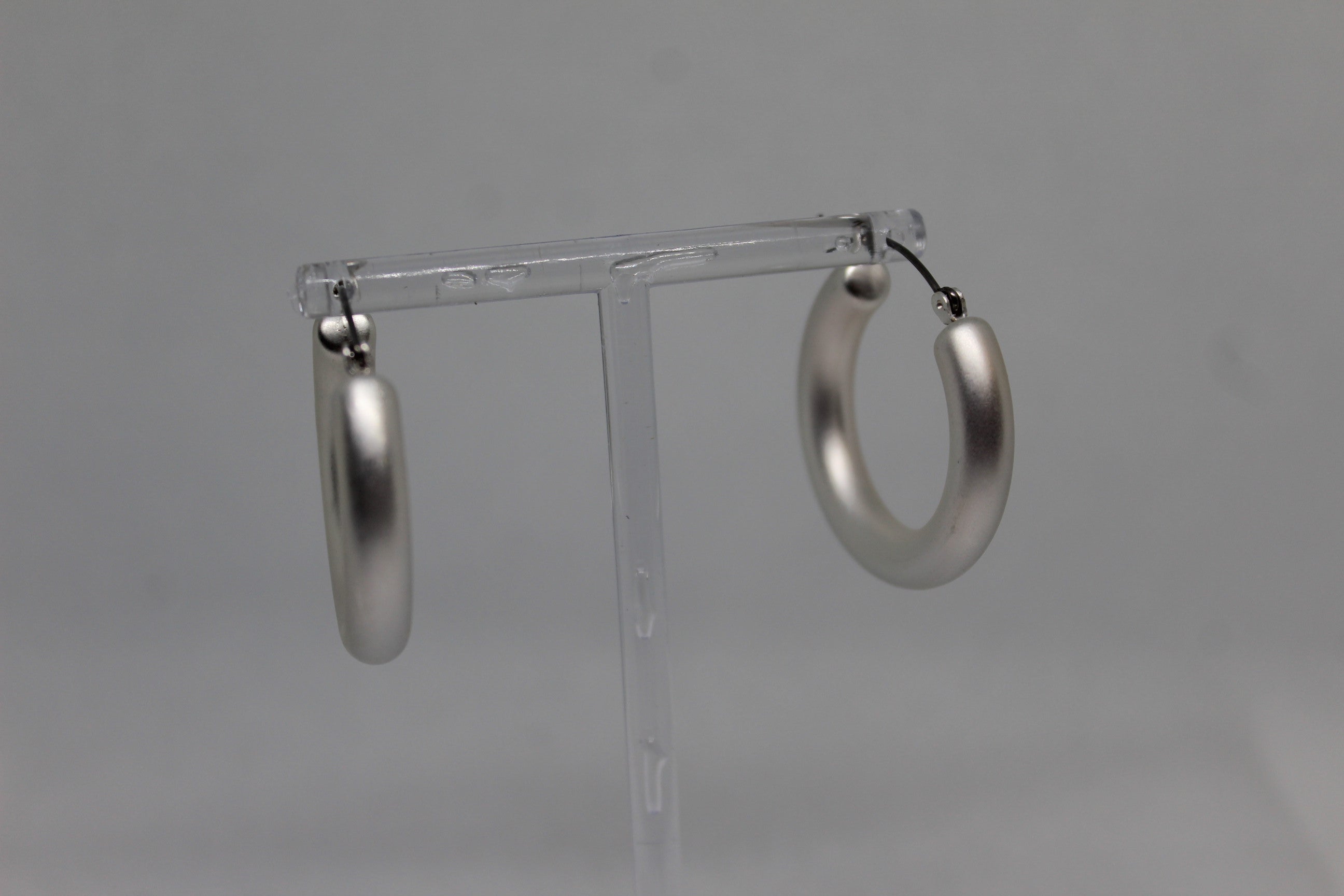 A pair of large, polished silver hoop earrings hanging from a clear plastic display stand against a neutral gray background.