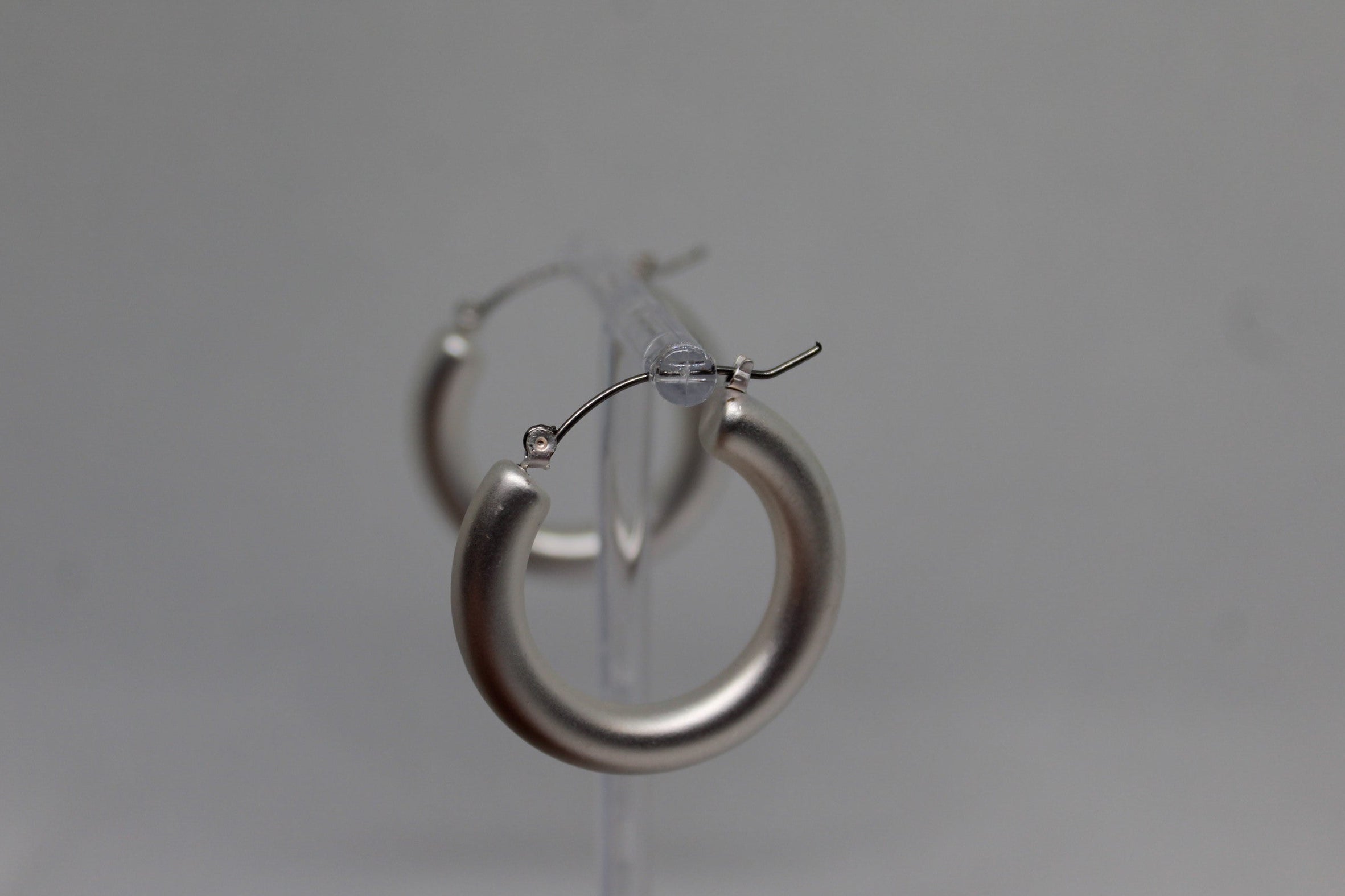 A pair of thick, polished silver hoop earrings displayed on a clear plastic stand against a neutral gray background.