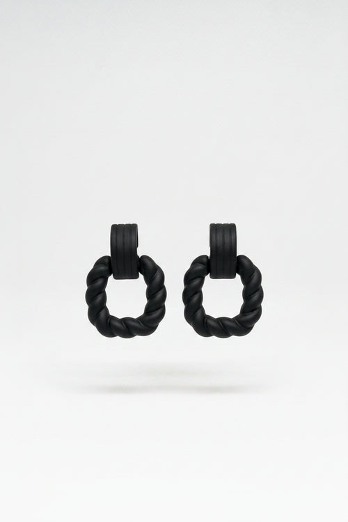 A pair of black braided hair ties with small wooden tags displaying the letters 'PLR', attached to a kraft paper display card with a white logo, set against a textured beige knitted background.