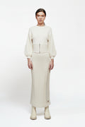 A model stands against a plain white studio background wearing a cream-colored knit two-piece outfit consisting of a long-sleeve top with a textured waistband and a matching long skirt, paired with beige ankle boots.