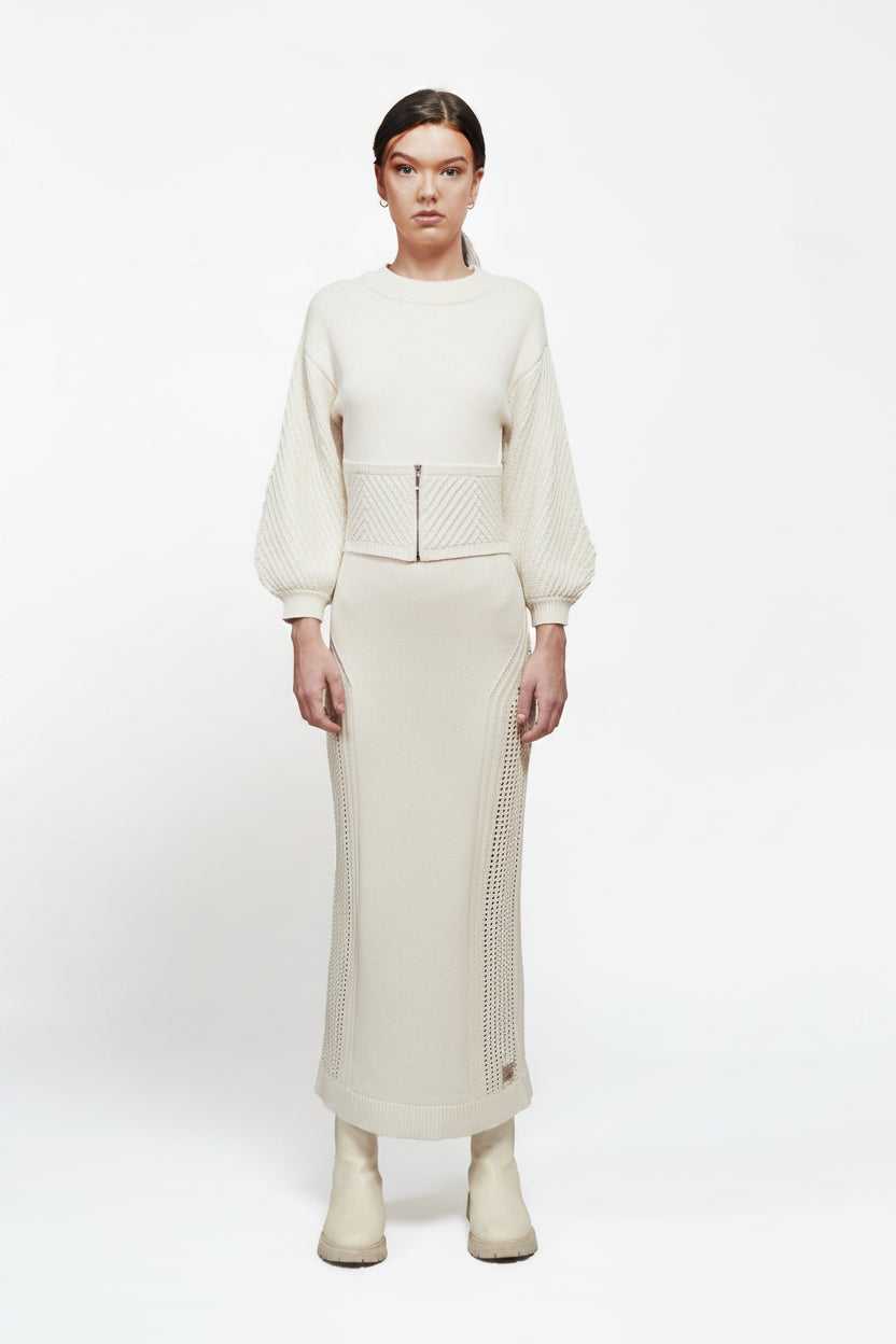 A model stands against a plain white studio background wearing a cream-colored knit two-piece outfit consisting of a long-sleeve top with a textured waistband and a matching long skirt, paired with beige ankle boots.