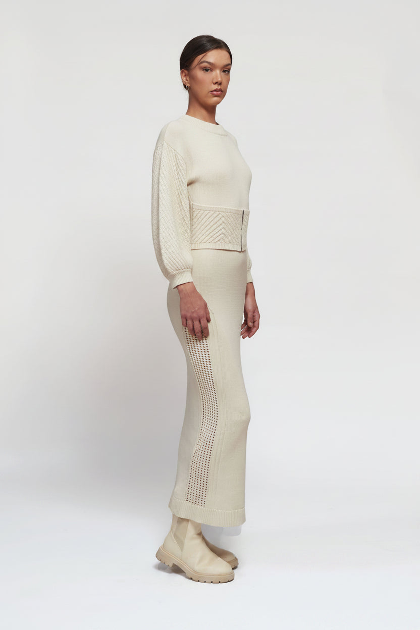 A model stands in profile wearing a cream-colored knit two-piece set consisting of a long-sleeve top with floral embroidery on the shoulder and a matching long skirt, paired with chunky cream ankle boots, against a plain white studio background.